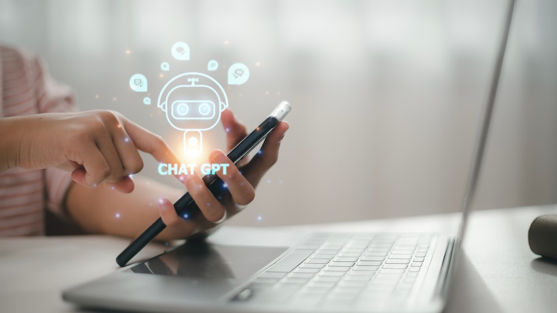 Handen (van een vrouw) toeten iets in op telefoon. Ook nog een laptop te zien. In een soort wolkje staat CHATGTP